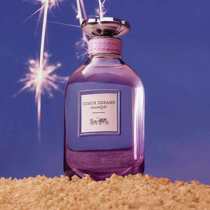 Purple Coach Dreams perfume bottle on sand with sparklers against a blue sky